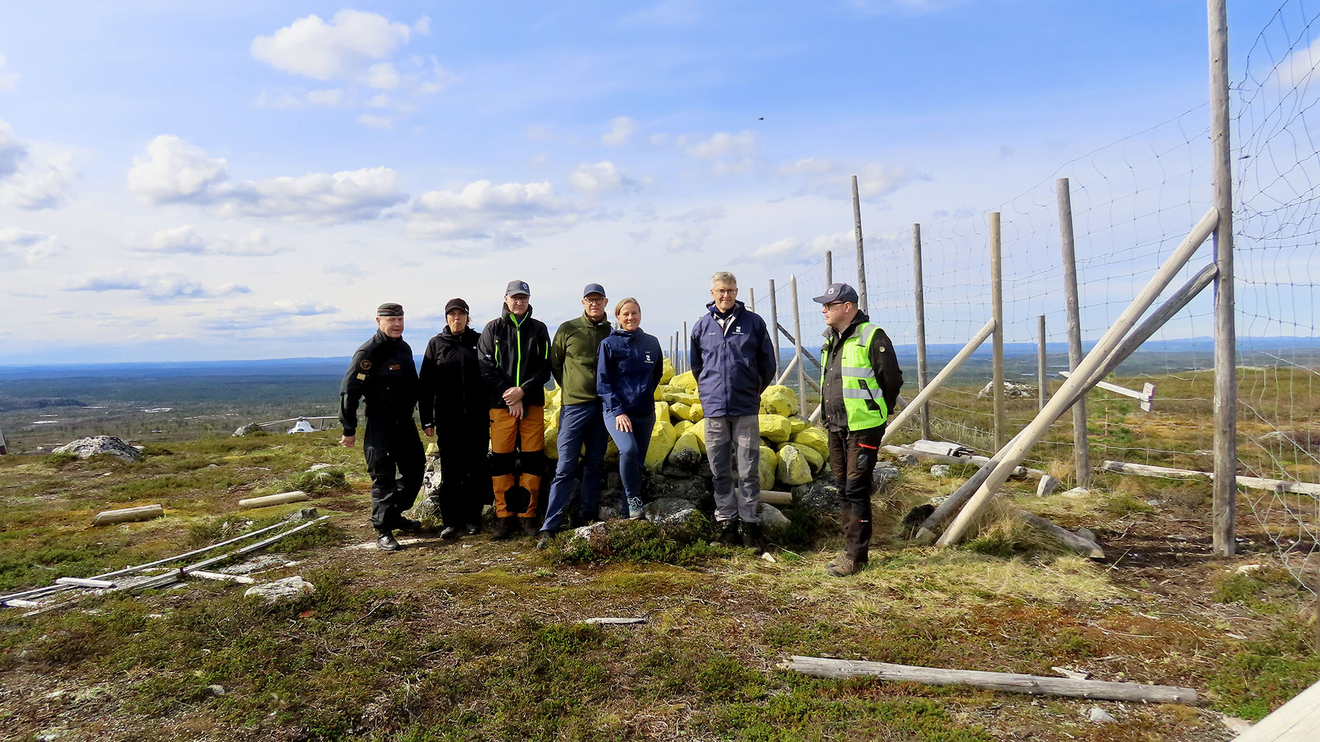 Seitsemän rajankäyntivaltuuskunnan jäsentä seisoo tundramaisemassa, oikealla puolella raja-aita.