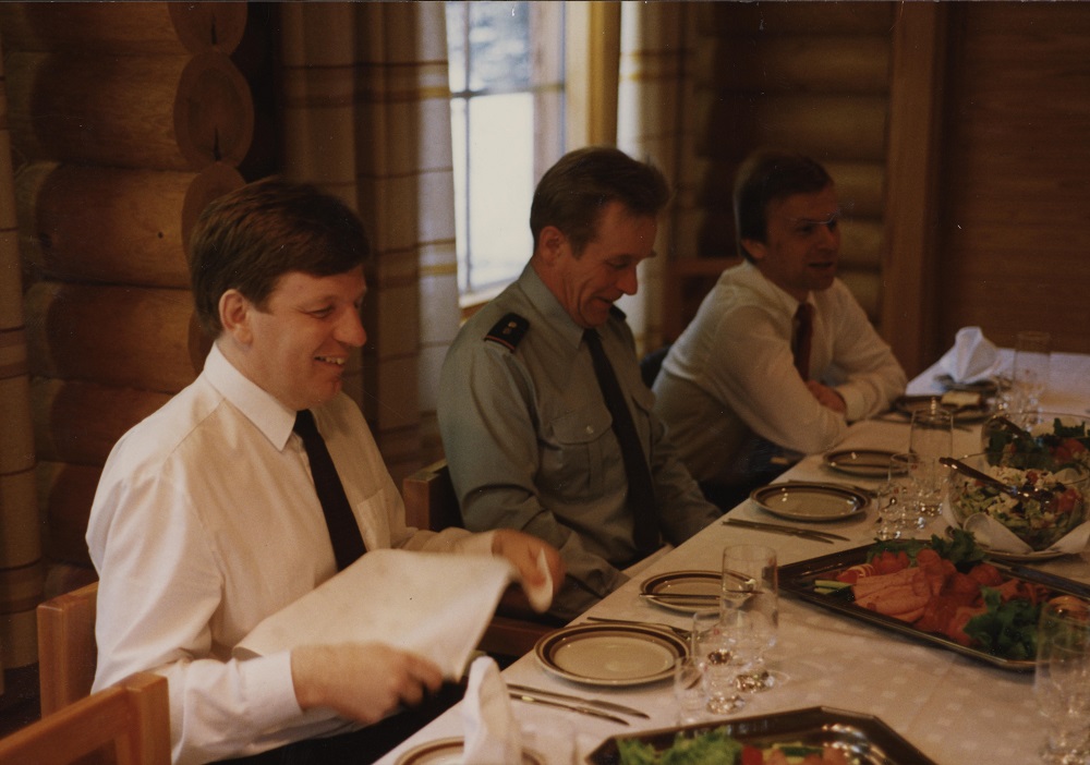 Männen ler och skrattar vid matbordet före måltiden. Statsminister Aho placerar servetten i famnen. Det finns sallad och skinkskivor på faten och i skålarna på bordet.
