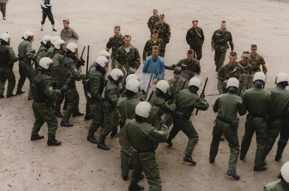 Toistakymmentä valmiusjoukkueen miestä harjoittelee hiekkakentällä mellakantorjuntaa pidellen käsissään suojakilpiä ja pamppuja. Miehiä vastassa on kymmenkunta maastopukuista miestä.