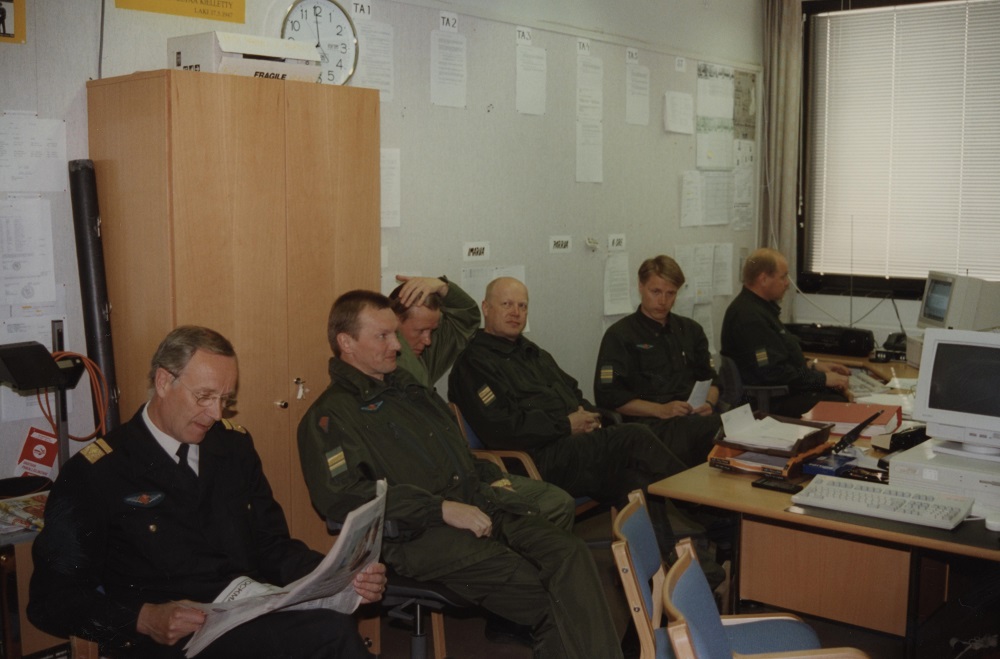 Men sit side by side in an office behind a desk looking expectant. An officer of the Finnish Coast Guard is reading a newspaper on the forefront.