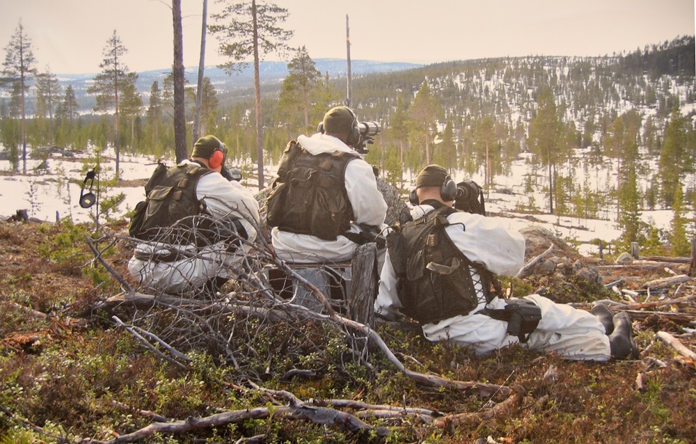 Men dressed in snow suits and combat vests use binoculars to monitor shrubby terrain in the winter. On the background is a sparsely forested fell landscape.