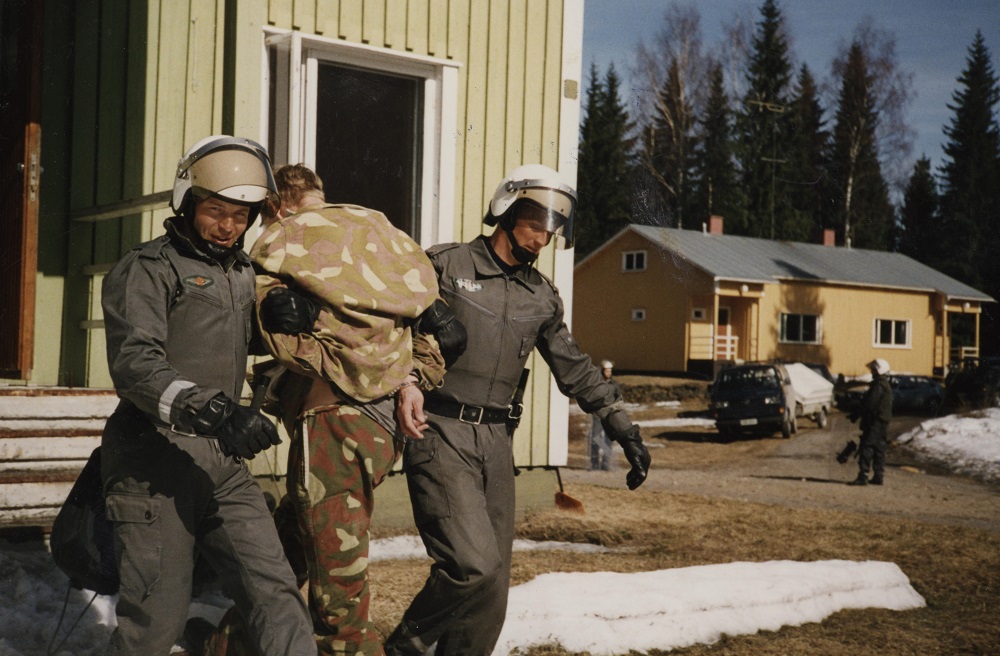 Two men wearing helmets and dressed in grey drag a man in a camouflage uniform from a wooden house. The man on the left has a small smile on his face.