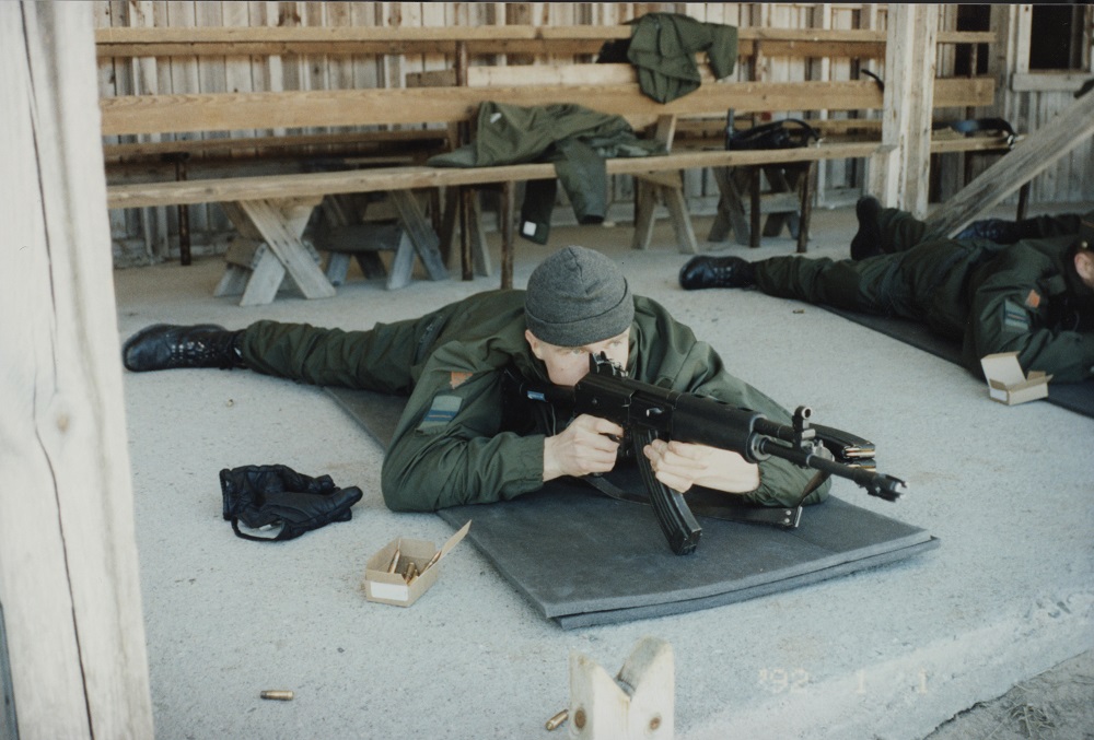 En man med mössa siktar med RK 65-stormgevär i liggande ställning på en tunn madrass. I närheten av mannens högra hand finns läderhandskar och en ask patroner.