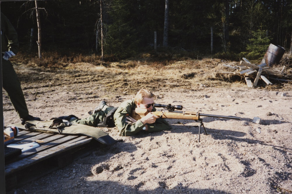 A man with sunglasses is lying down and aiming with a TKIV 85 sniper rifle in sandy terrain. Behind him is a man standing with his legs wide apart who is watching the hits with the rifle.