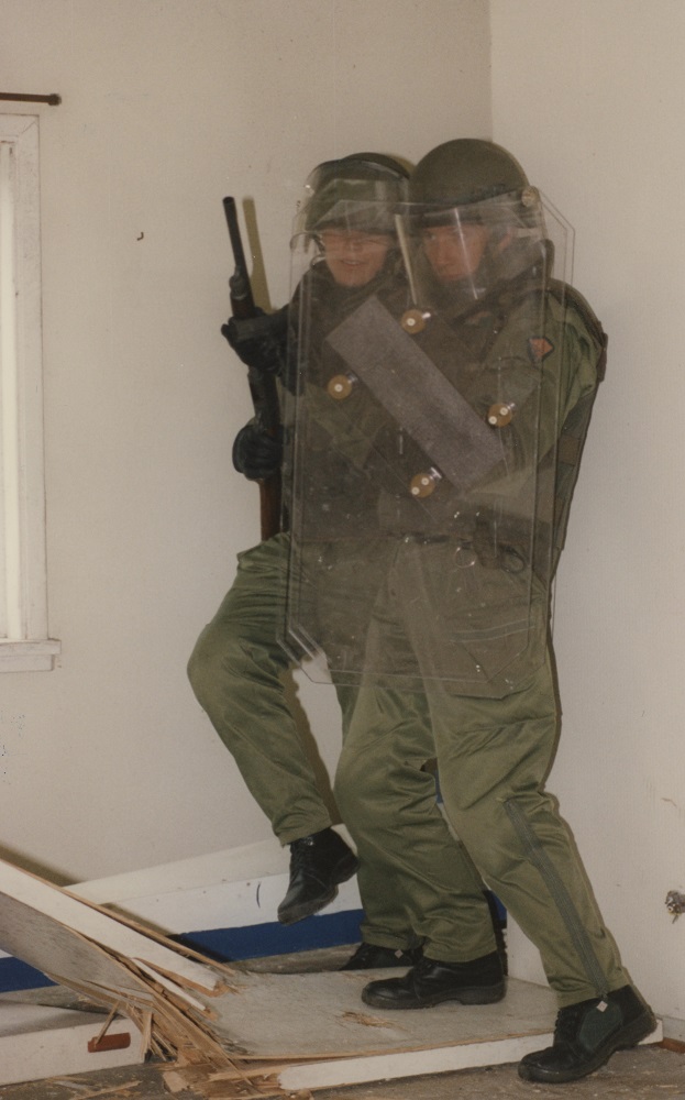 Två män med skyddsutrustning inne i byggnaden framför väggen. Den främre mannen håller en skyddssköld. Den bakre mannen har ett hagelgevär.