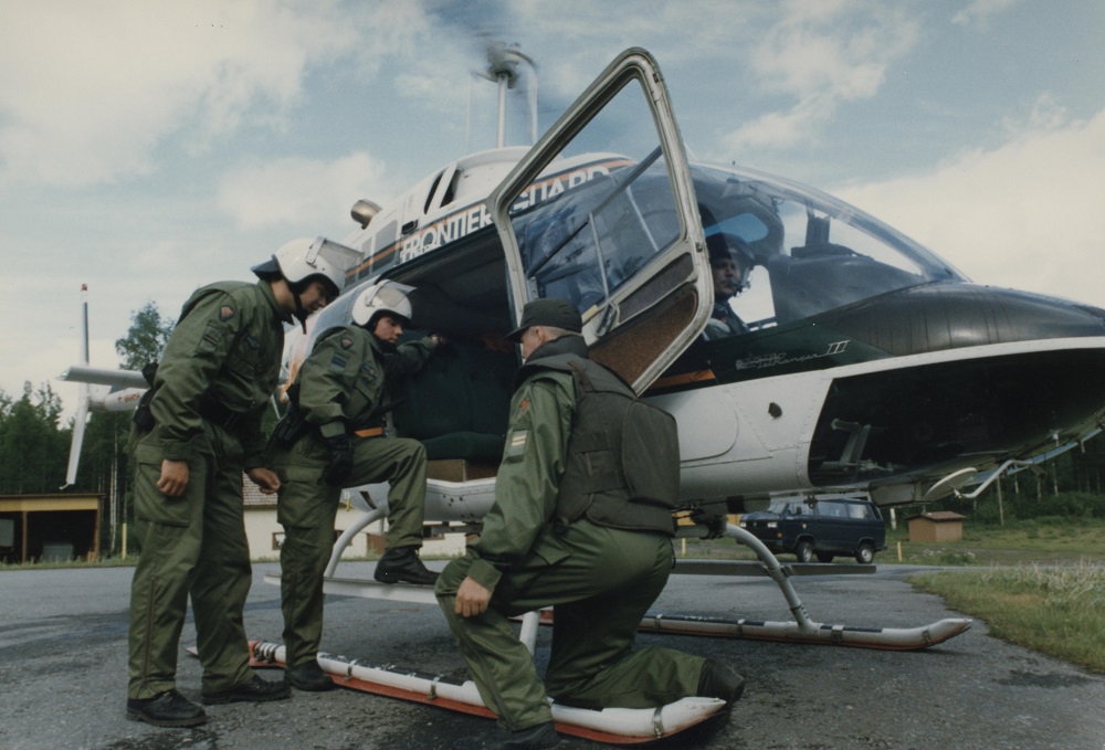 Two men wearing helmets are boarding a running Border Guard helicopter. A man in a cap in front of them is looking at the men boarding the helicopter. The pilot of the helicopter is visible in the cockpit and is looking towards the camera.