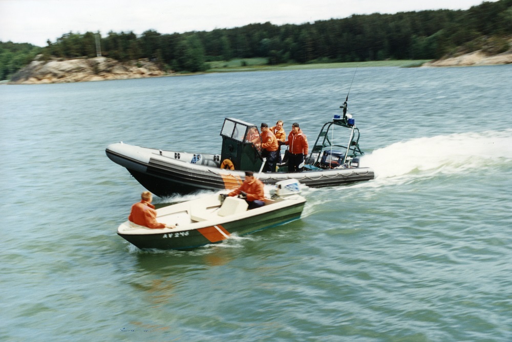 Men wearing orange shirts in two boats in the water. The speedboat behind them is moving fast and passing the small boat. A forest and rocky beach are visible on the background.