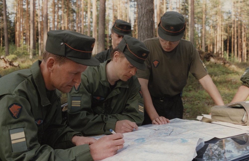 Men in a forest are leaning onto a topographic map and looking at it. The front two men in long sleeved uniforms are pointing and marking the map with a pen.