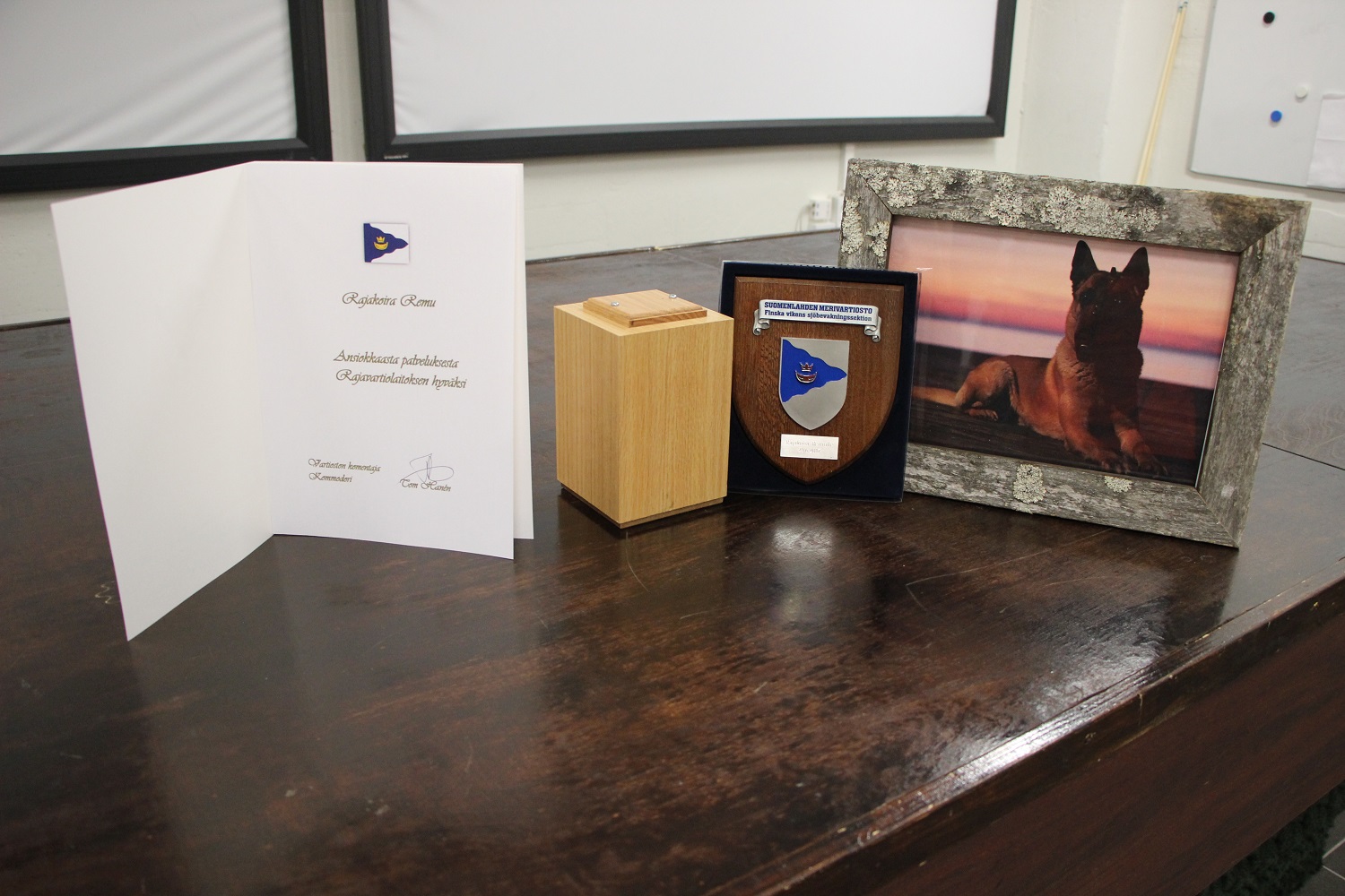 A memorial card, a wooden object, a coat of arms of the Gulf of Finland Coast Guard District and a framed photo of Remu as a puppy are on the table.
