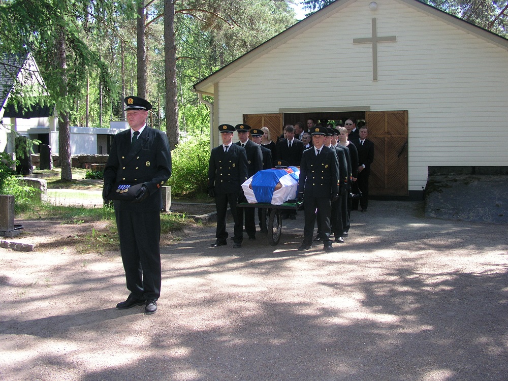 Men wearing dark clothes carry a coffin wrapped in the Finnish flag in front of a chapel building on a summer day. In the foreground, a man is holding medals in his hands.