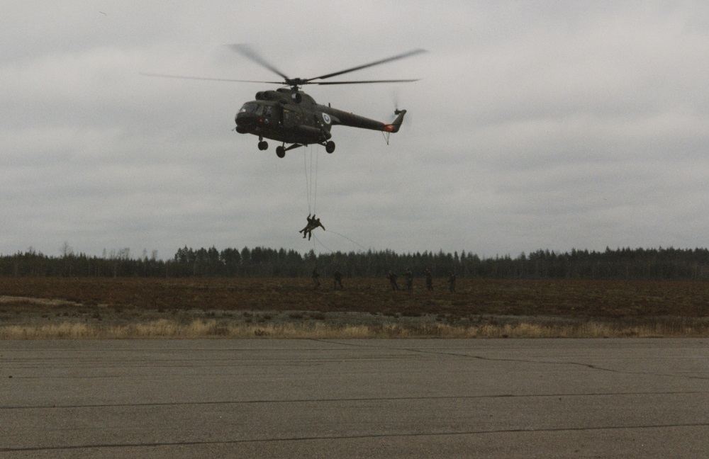 Two people hang from ropes from an airborne helicopter. Under the helicopter, six men from the special intervention unit are visible on the ground.