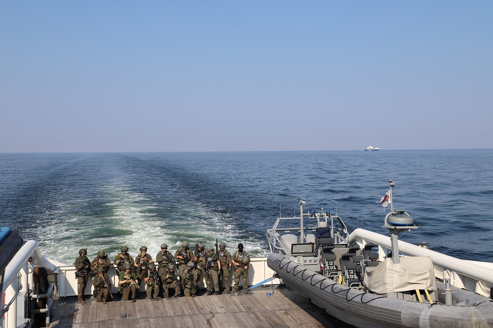 En grupp i Gränsbevakningsväsendets beredskapspluton och polisens Beredskapsenhet Karhu poserar i rad bak på fartyget. I bakgrunden ett öppet hav och några fartyg i horisonten. Det finns en snabbåt bredvid männen.