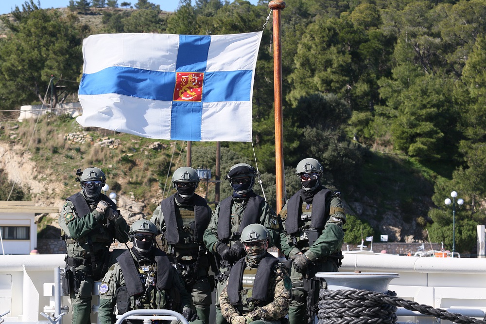 The operators from the special intervention unit are wearing ski masks and helmets and posing for a photo under the Finnish flag on a ship's deck. Forest terrain and harbour area can be seen in the background.