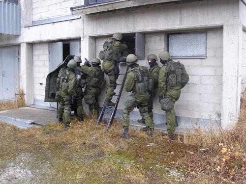 En grupp män från beredskapsplutonen framför väggen på ett flervåningshus. En av männen är på en stege och siktar in med sin pistol genom ett öppet fönster.