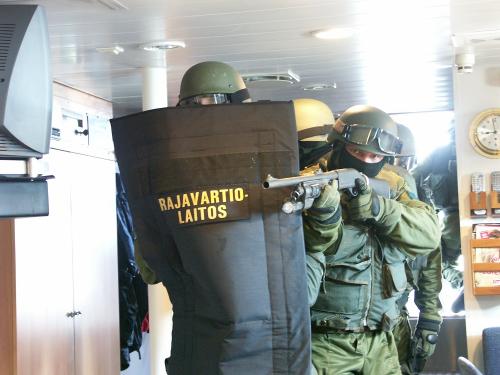 A group of men of the special intervention unit are walking towards the photographer inside of a building or a ship. One man is holding an assault shield. Another man takes an aim with his shotgun.
