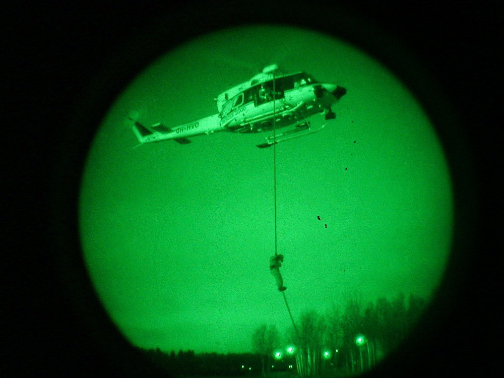 A view seen through night vision goggles shows a descending man from a helicopter via a rope.