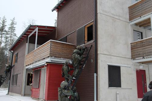 Three men of the special intervention unit are climbing the ladders towards a window by a building. Two men are covering them.