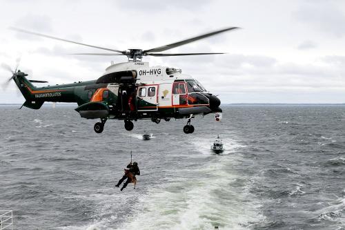 A helicopter hovers over water. It is winching down one man and a dog. A boat approaches from behind.