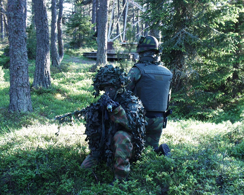 Kaksi maastopukuista valmiusjoukkueen operaattoria polvillaan metsässä. Edessä oleva mies osoittaa aseella. Selin oleva mies tähystää taaksepäin.