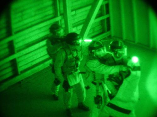 Fyra av beredskapsplutonens operatörer avancerar i en mörk byggnad. Den främre mannen skyddar de andra med en skyddssköld. De andra männen är redo att använda sina vapen.