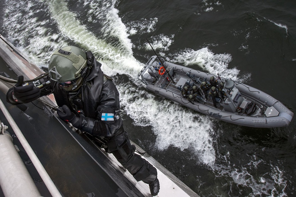 En beredskapsman klädd i mörk skyddsdräkt och skyddsutrustning klättrar på en stege upp längs sidan av en båt. Under mannen finns en gummibåt från vilken han har kommit upp på stegen. Det är sju man ombord.
