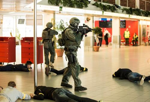 Två beväpnade grönklädda operatörer från beredskapsplutonen avancerar i hamnterminalbyggnaden. Fem personer ligger på golvet. I bakgrunden syns människor som följer övningen.