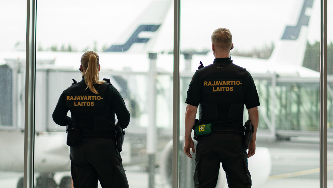 Kaksi rajavartijaa katsoo lentoaseman ikkunasta selät kameraan päin.
