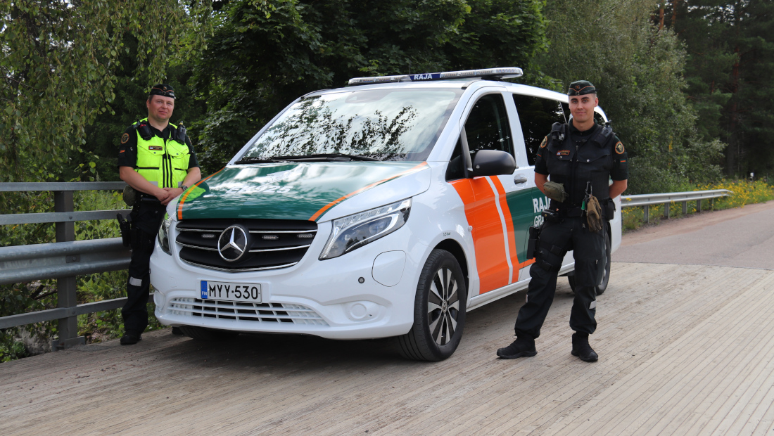 Kaksi rajavartijaa seisoo partioauton vierellä puisella sillalla.