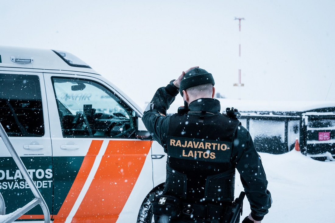 Rajavartija seisoo selkä kohti kameraa partioauton vieressä lumisella lentokentällä.