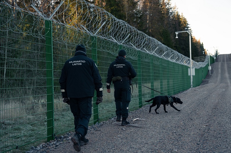 Two border guards and a border guard dog are on the road next to the barrier fence.