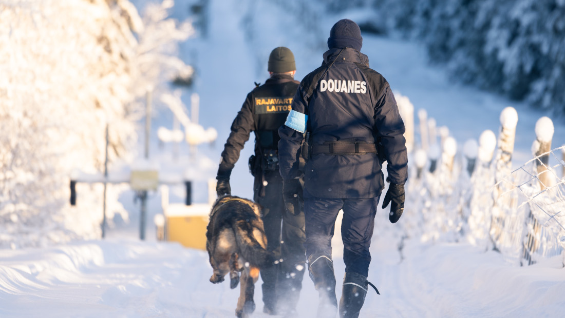 Frontex och Finlands gränsbevakare rör sig vid gränsen i snö bort från kameran. Hunden springer bredvid.