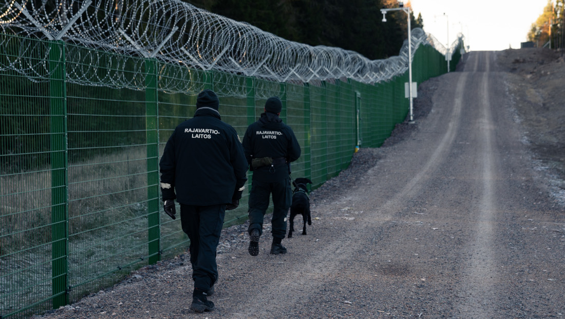 Two border guards and a border dog are walking next to the border barrier fence.