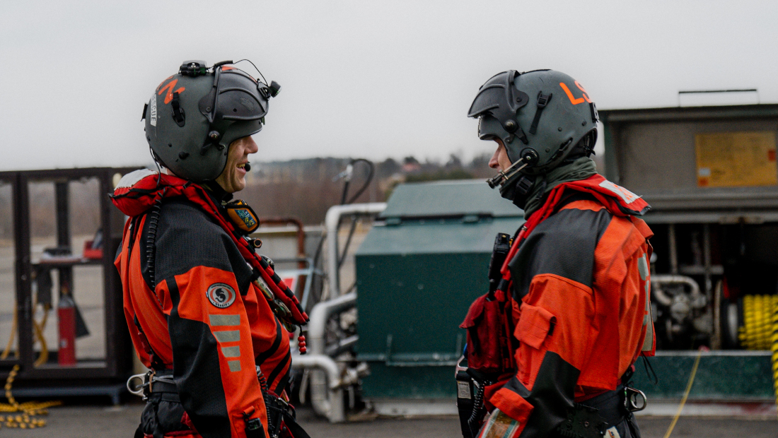 Two people in protective clothing are talking. Oil spill response equipment in the background.