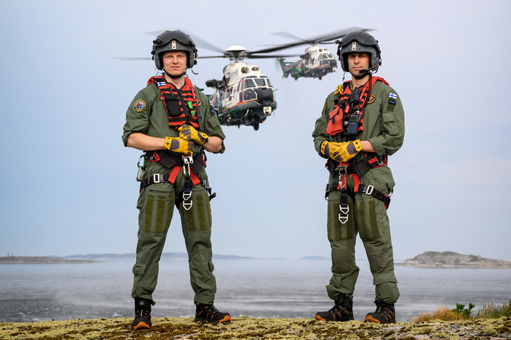 Två helikopterpiloter står på en kobbe. I bakgrunder havsvy och två helikoptrar i luften.