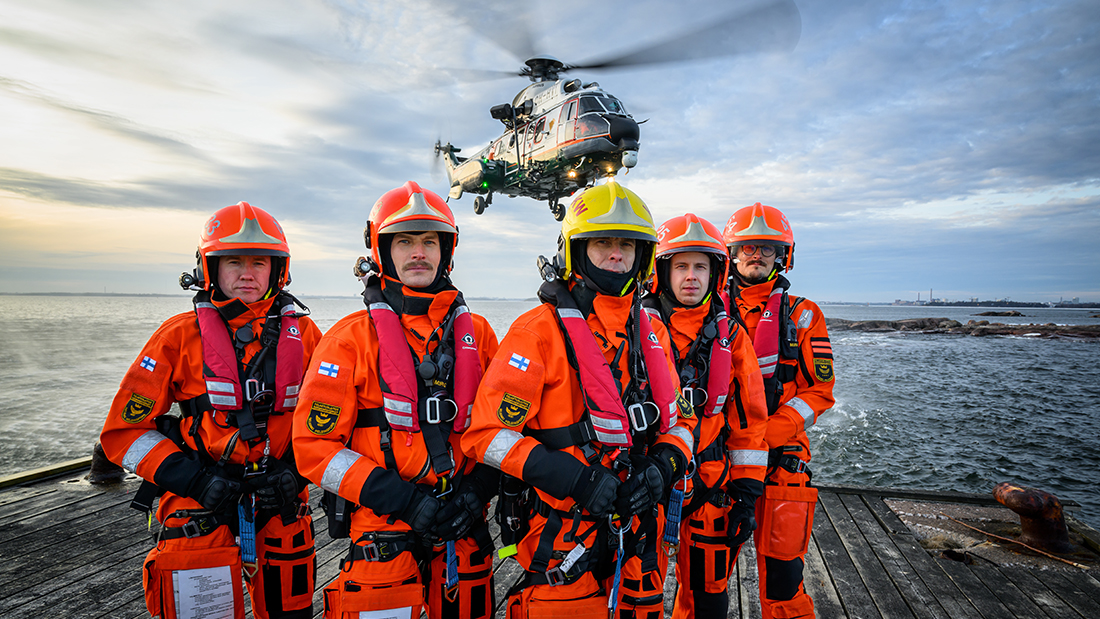Viisi pintapelastajaa seisoo rannalla. Takana merimaisema ja helikopteri ilmassa.