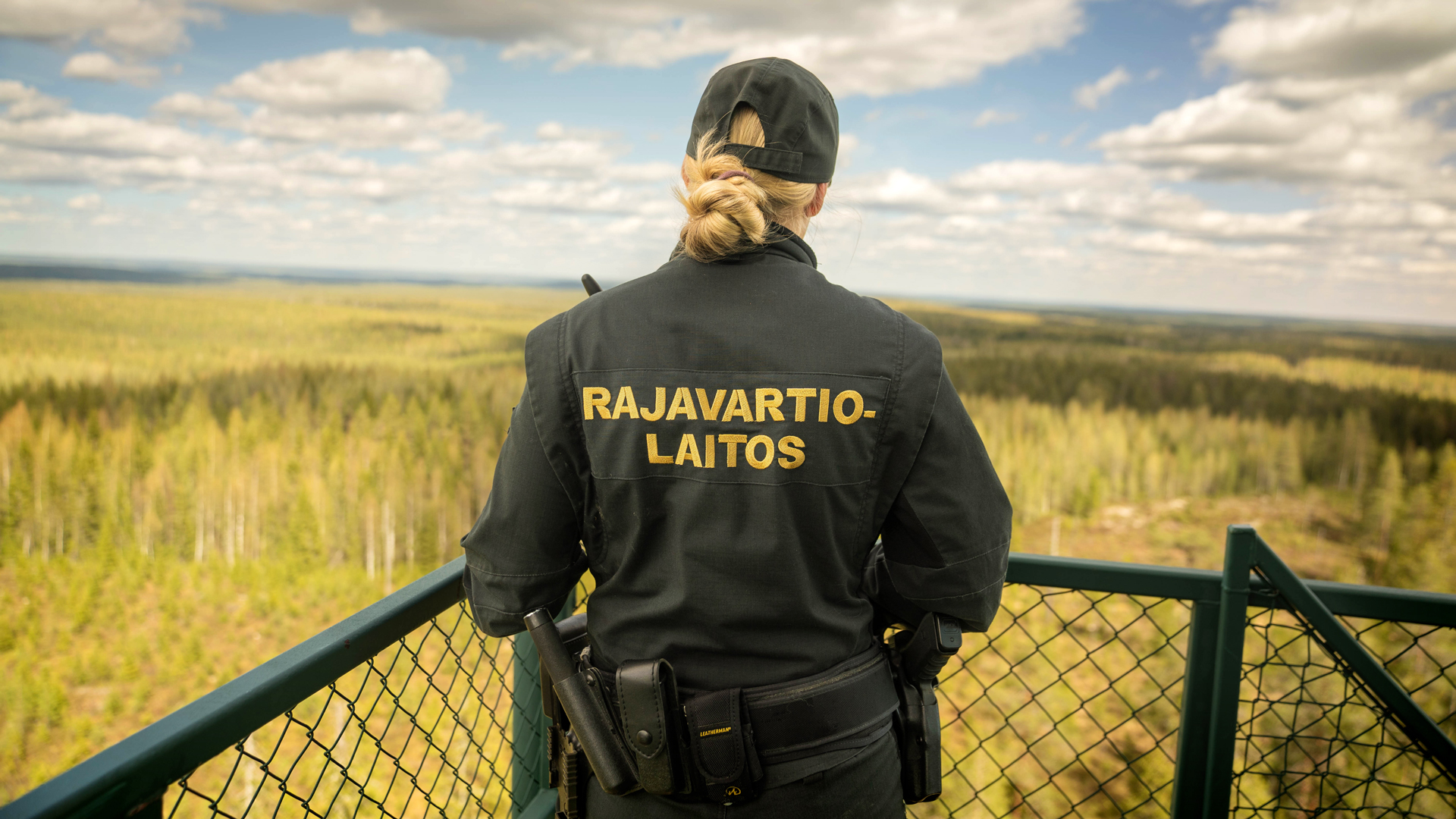 Gränsbevakaren står i vakttornet med ryggen mot kameran och tittar på det somriga åkerlandskapet framför sig.