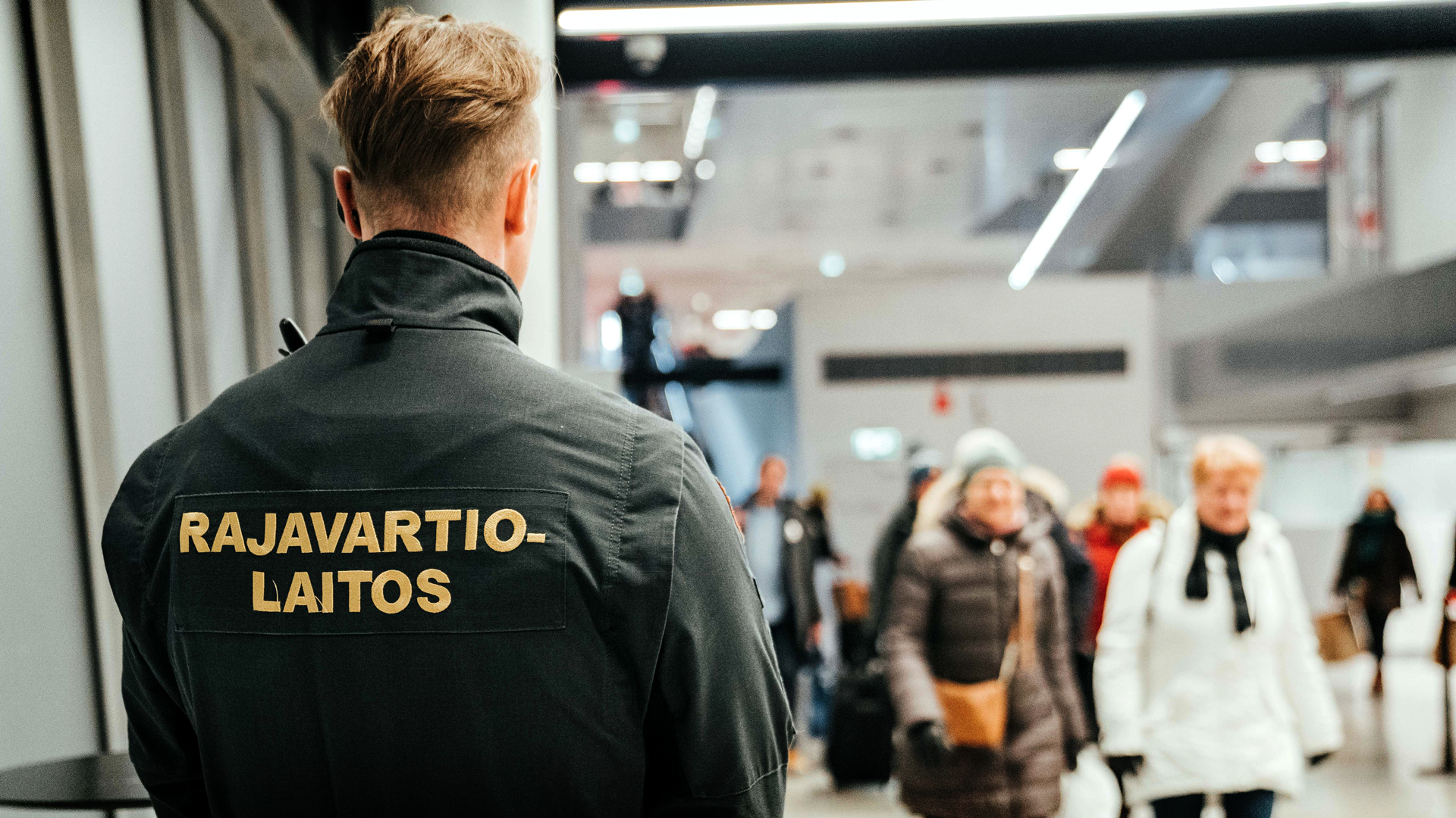 A border guard standing on the airport facing away from the camera, overseeing passengers passing by.