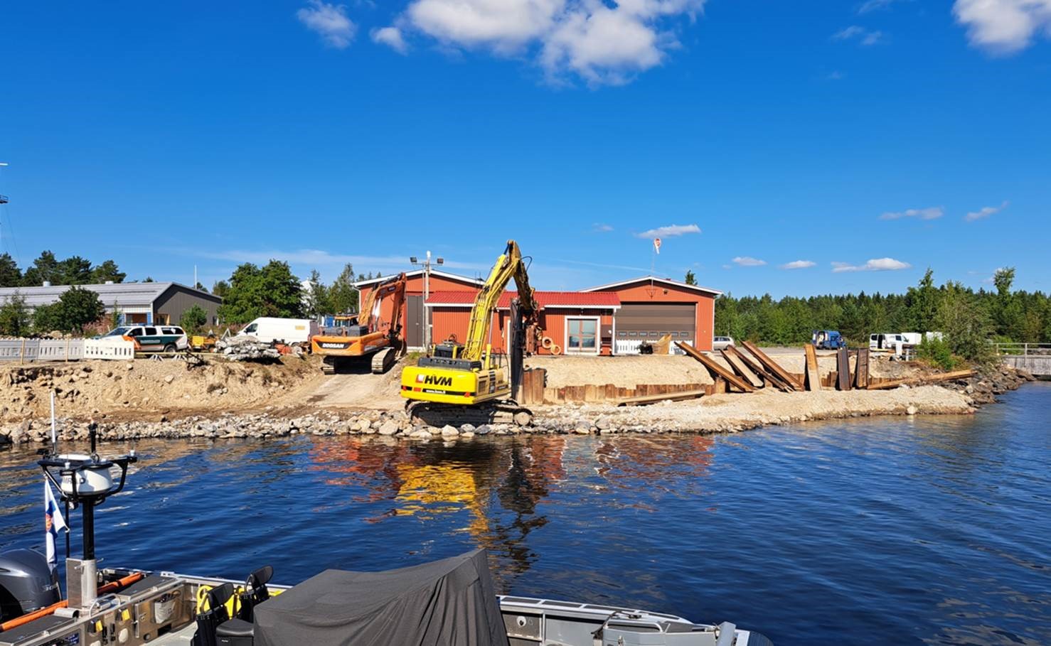 Virpiniemi Coast Guard Station under construction. An excavator on the beach with wooden buildings, painted red, in the background.