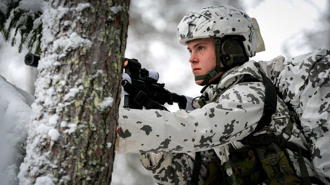 Halvfigursporträtt av en gränsvakt i vinterterräng. Gränsvakten bär vinterkläder och håller ett vapen, delvis bakom ett träd.