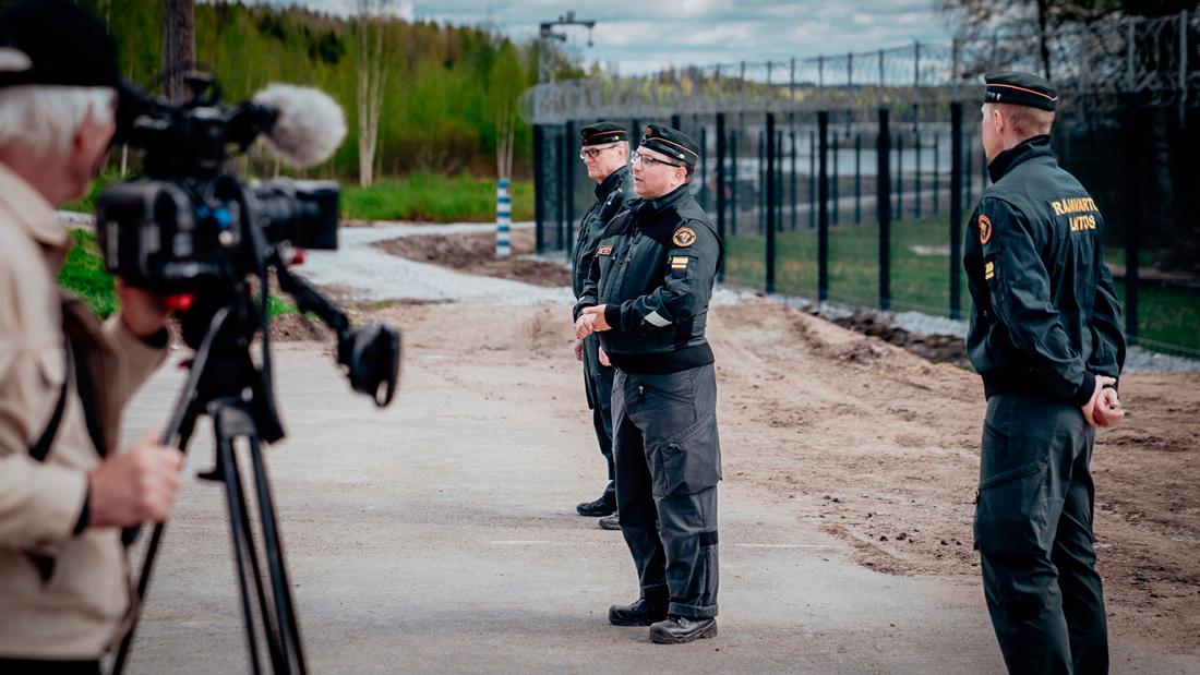 En gränsbevakningsofficer står framför det östra stängslet och talar med medierepresentanter. Fotografer och reportrar i förgrunden. Sommarlandskap i bakgrunden.