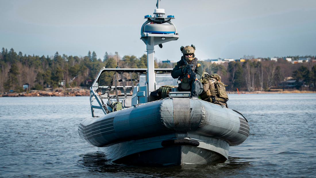 En medlem av gränsbevakningens räddningsstyrka står vid fören på en motorbåt vänd mot kameran i soligt sommarväder.