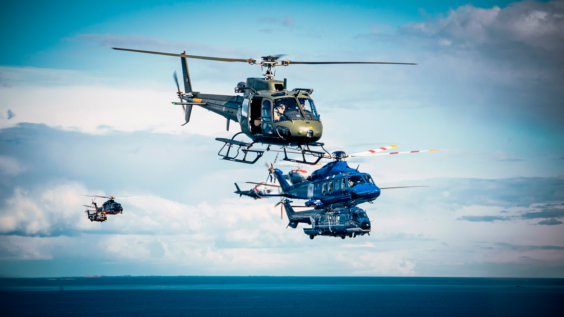 På bilden flyger tre helikoptrar i olika färger i luften i soligt väder. Vid horisonten syns ytterligare två helikoptrar. Nedanför, det blå havet.