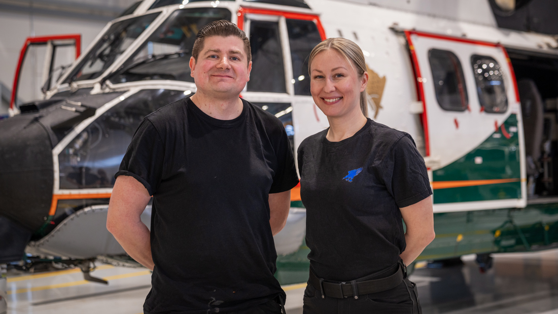 En man och en kvinna står i en helikopterhangar och tittar leende in i kameran. En gränsbevakningshelikopter syns i bakgrunden.