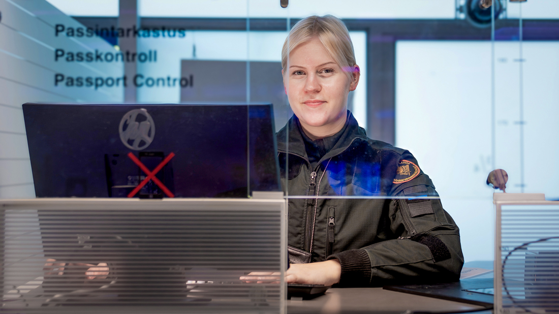 En ung kvinnlig gränsvakt sitter i en passkontrollbås och tittar in i kameran.