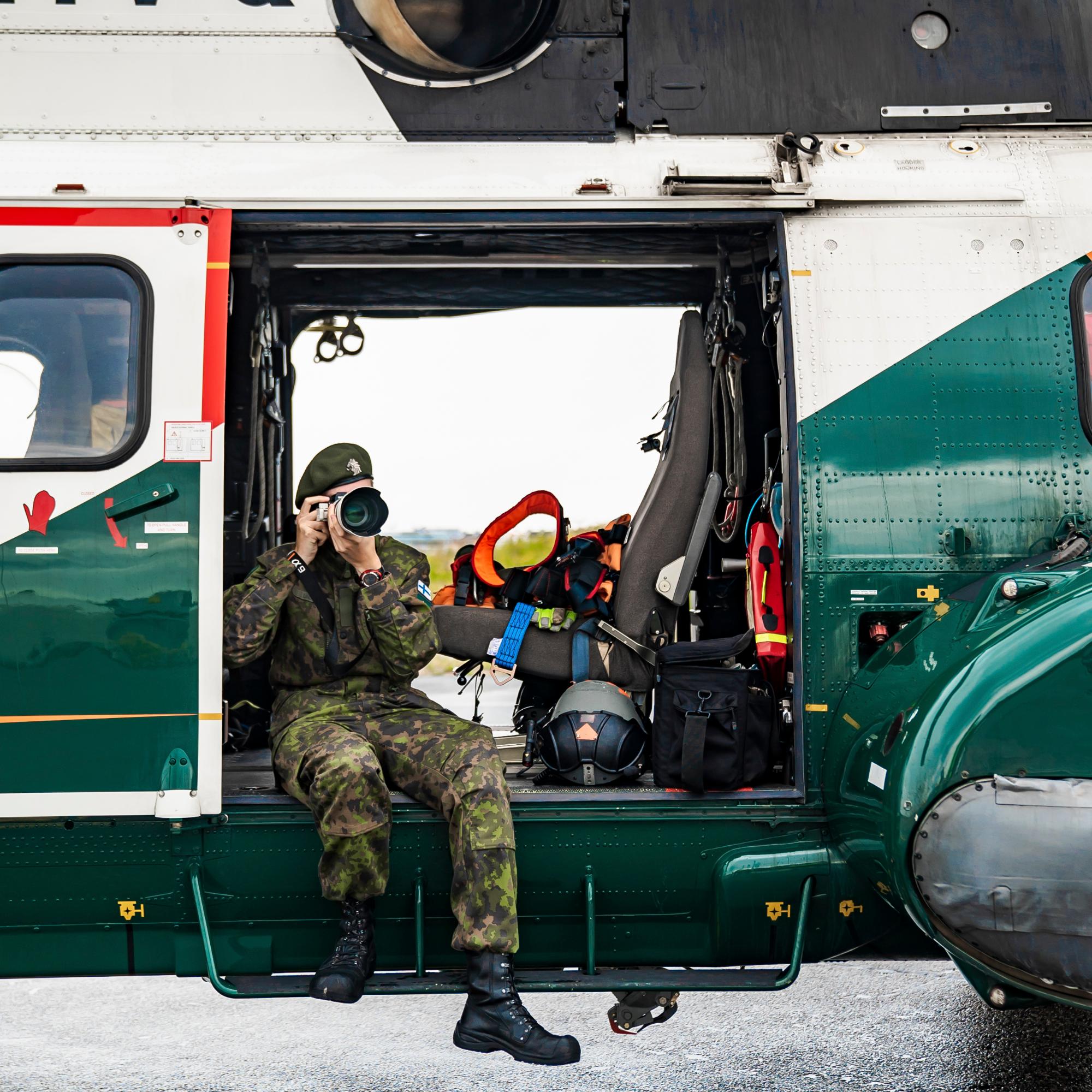 Varusmies istuu helikopterin oviaukolla ja ottaa valokuvaa.
