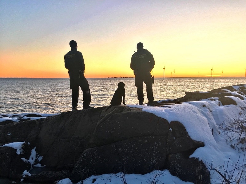 Två figurer står på en delvis snöig klippa med ryggen mot fotografen, mellan dem sitter en hund. De tittar på solnedgången vid havet, vid horisonten syns vindkraftverk.