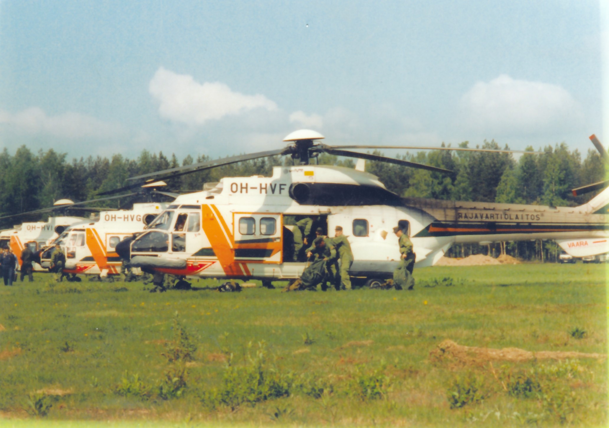 Three helicopters have landed on a grass field. A group of men are unloading equipment from the foremost helicopter.