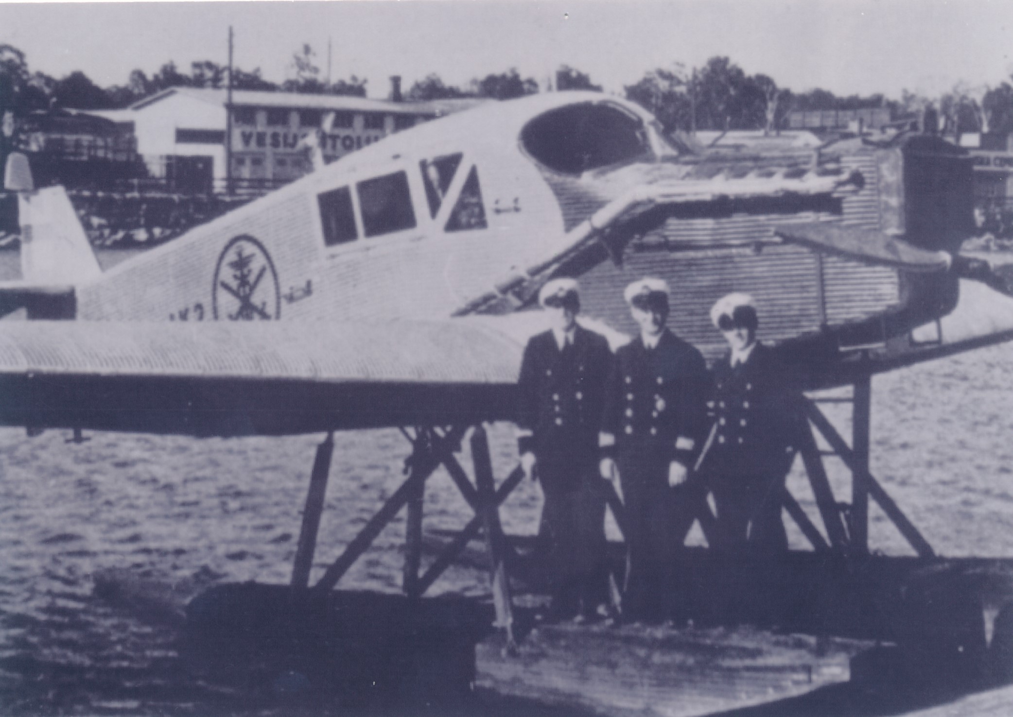 A seaplane, fitted with metallic floats, floating in the water. A uniformed, three-person crew is standing on one of the floats.