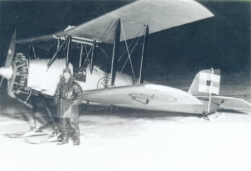 A biplane fitted with snow landing gear in a snow-filled landscape. The pilot standing in front of the aircraft is dressed in old-fashioned, leather flying gear.
