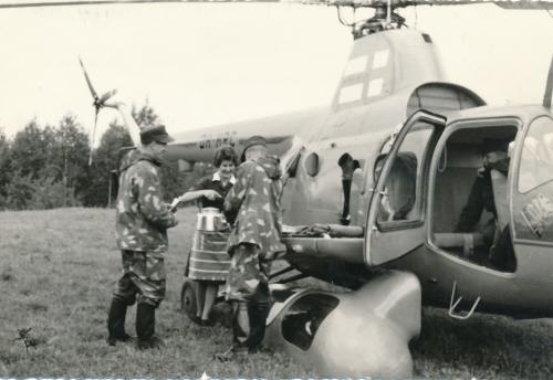 A helicopter has landed in a field. In front of the helicopter are two soldiers wearing camouflage. One of the green sisters is serving the soldiers from a coffee kettle.